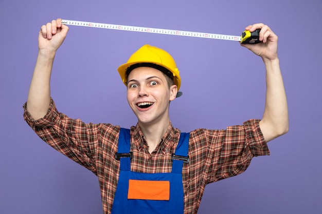 Macho joven constructor vistiendo uniforme que se extiende cinta métrica aislada en la pared púrpura