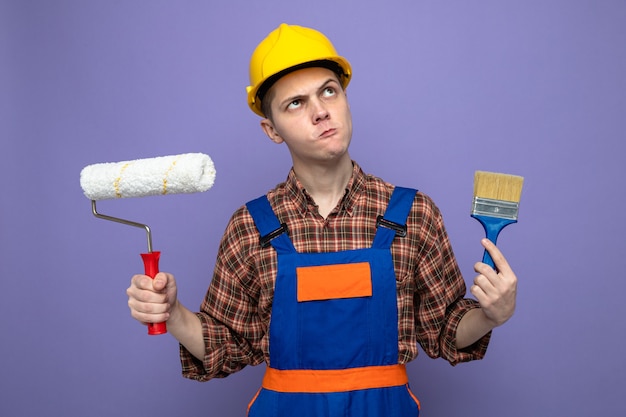 Macho joven constructor vistiendo uniforme sosteniendo pincel con cepillo de rodillo aislado en la pared púrpura