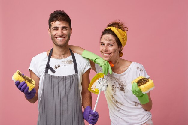 Macho joven con delantal con ropa sucia sonriendo ampliamente sosteniendo una esponja y cepillo y una mujer bonita inclinada en su hombro sosteniendo una esponja y detergente feliz de terminar la limpieza de primavera