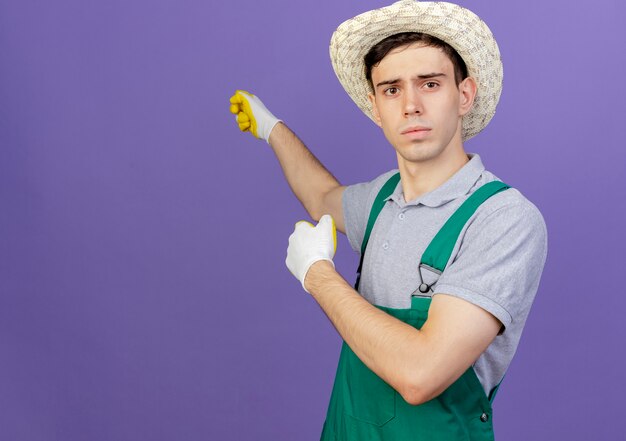 Macho joven desorientado jardinero vistiendo guantes y sombrero de jardinería puntos detrás