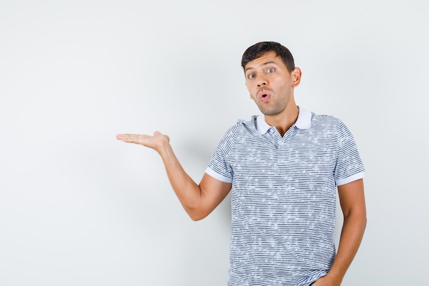 Macho joven extendiendo la palma a un lado en camiseta y mirando sorprendido