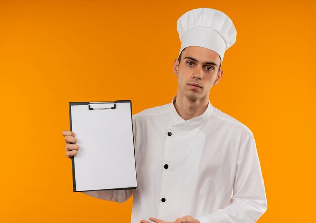 Macho joven fresco vistiendo uniforme de chef mostrando el portapapeles en su mano en la pared amarilla aislada