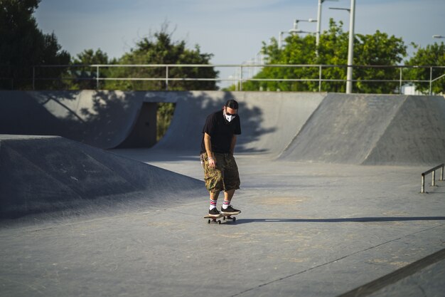 Macho joven haciendo diferentes trucos con una patineta en el parque con una mascarilla médica