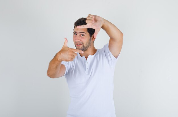 Macho joven haciendo gesto de marco en camiseta blanca y mirando feliz