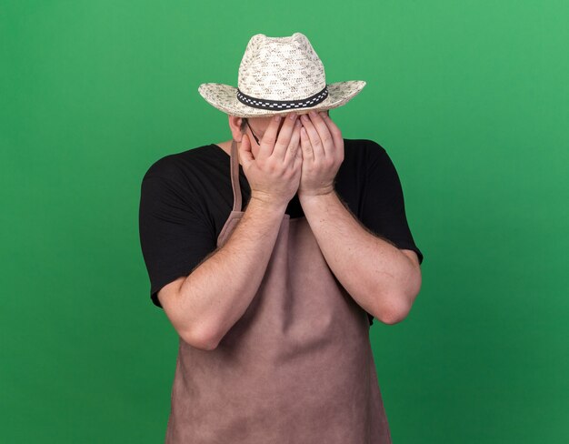 Macho joven jardinero arrepentido con sombrero de jardinería rostro cubierto con las manos aisladas en la pared verde