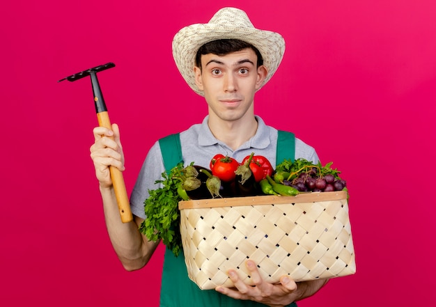 Foto gratuita macho joven jardinero complacido vistiendo sombrero de jardinería tiene canasta de verduras y rastrillo