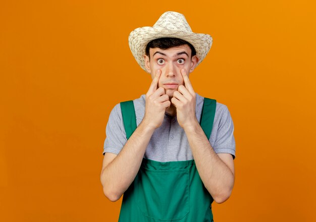 Macho joven jardinero conmocionado vistiendo sombrero de jardinería tira hacia abajo los párpados