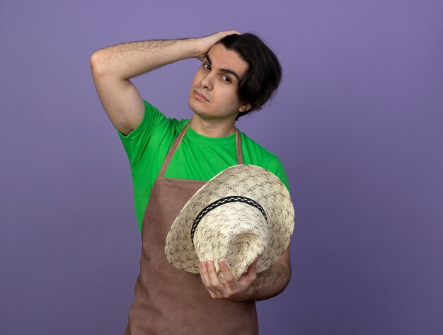 Foto gratuita macho joven jardinero en uniforme sosteniendo sombrero de jardinería poniendo la mano en la cabeza.
