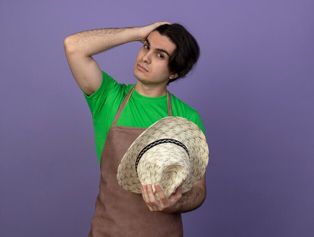 Macho joven jardinero en uniforme sosteniendo sombrero de jardinería poniendo la mano en la cabeza.