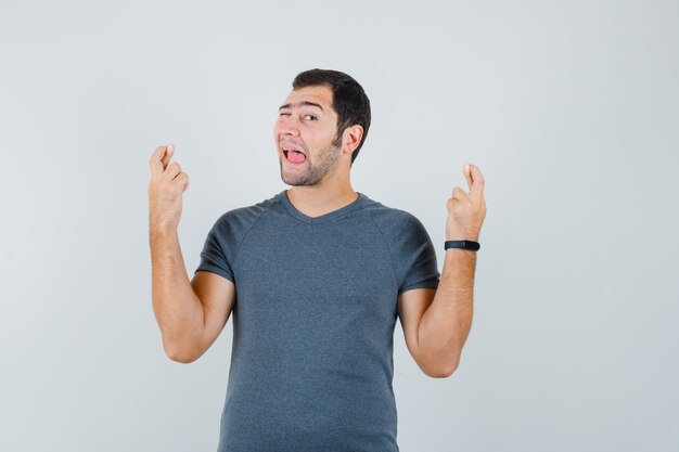 Macho joven manteniendo los dedos cruzados guiñando el ojo sacando la lengua en camiseta gris