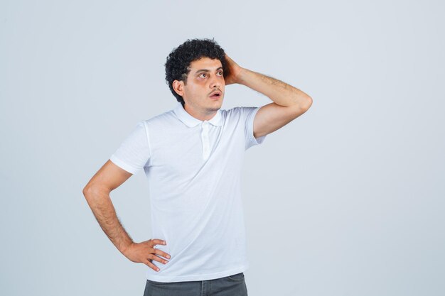 Macho joven manteniendo la mano en la cabeza en camiseta blanca, pantalones y mirando sorprendido, vista frontal.