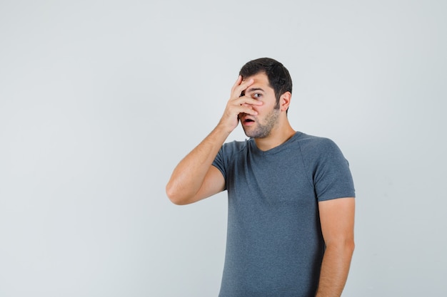 Macho joven mirando a través de los dedos en camiseta gris y mirando asustado