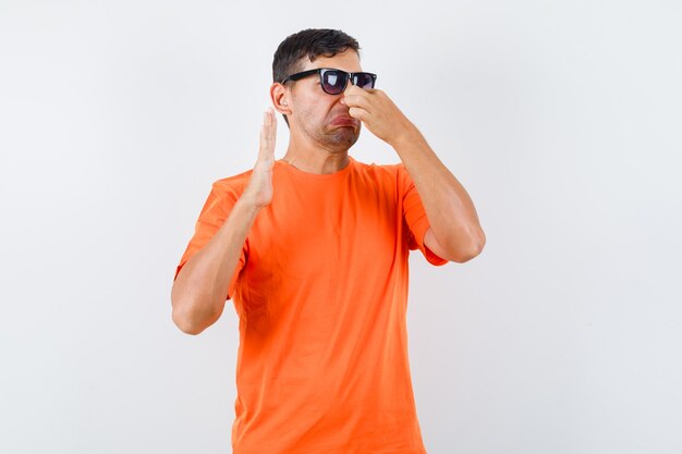 Macho joven pellizcando la nariz debido al mal olor en camiseta naranja y mirando disgustado