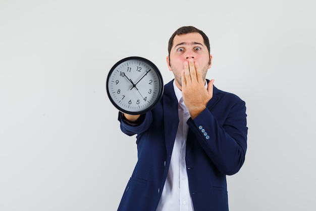 Macho joven que muestra el reloj de pared en camisa y chaqueta y mirando asustado