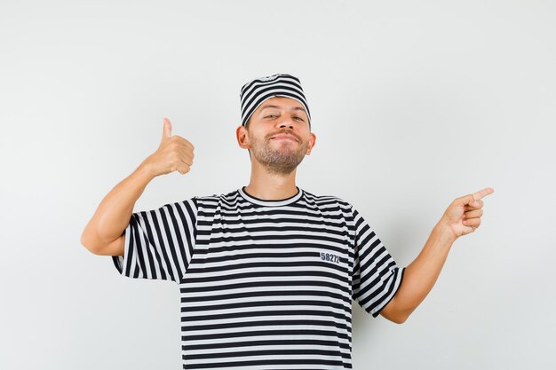 Macho joven con sombrero de camiseta a rayas apuntando hacia el lado mostrando el pulgar hacia arriba y mirando alegre
