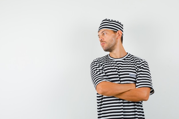 Macho joven con sombrero de camiseta a rayas mirando a un lado con los brazos cruzados y mirando enfocado