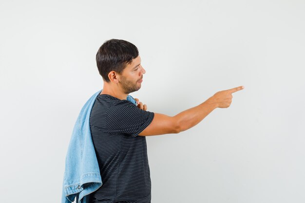 Foto gratuita macho joven sosteniendo la chaqueta en la espalda apuntando hacia afuera en camiseta y mirando positivo