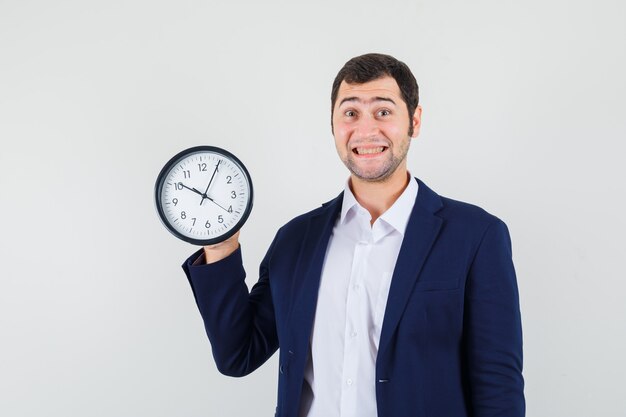 Macho joven sosteniendo el reloj de pared en camisa y chaqueta y mirando alegre