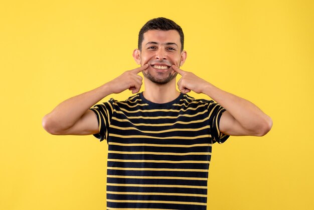 Macho joven de vista frontal en camiseta a rayas blanco y negro apuntando a su sonrisa sobre fondo amarillo aislado