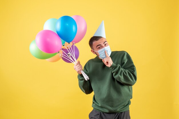 Macho joven de vista frontal sosteniendo globos de colores en el escritorio amarillo