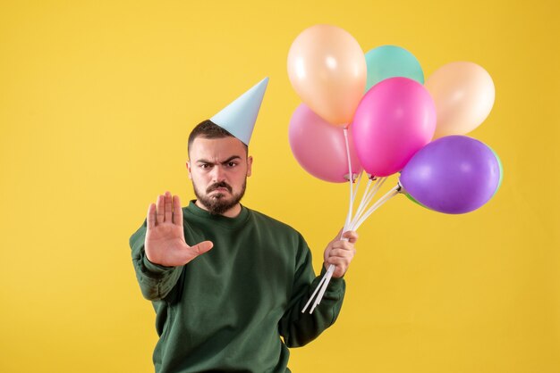 Macho joven de vista frontal sosteniendo globos de colores en el escritorio amarillo