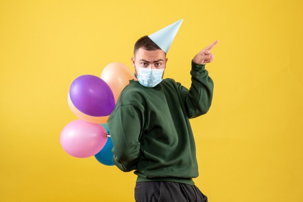 Macho joven de vista frontal sosteniendo globos de colores sobre un fondo amarillo