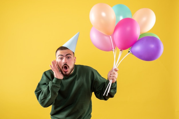 Macho joven de vista frontal sosteniendo globos de colores sobre un fondo amarillo