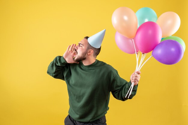 Macho joven de vista frontal sosteniendo globos de colores sobre un fondo amarillo