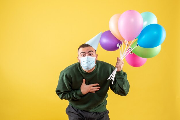 Macho joven de vista frontal sosteniendo globos de colores sobre un fondo amarillo