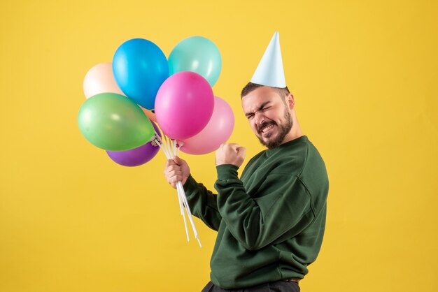 Macho joven de vista frontal sosteniendo globos de colores sobre un fondo amarillo