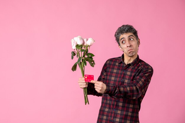 Macho joven de vista frontal sosteniendo hermosas rosas rosadas y tarjeta bancaria en la pared rosa