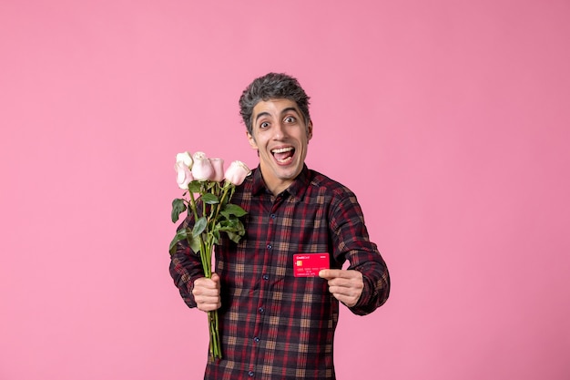 Macho joven de vista frontal sosteniendo hermosas rosas rosadas y tarjeta bancaria en la pared rosa