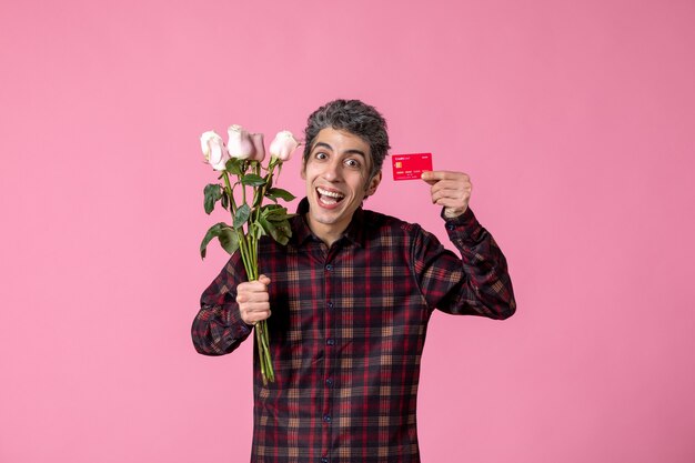 Macho joven de vista frontal sosteniendo hermosas rosas rosadas y tarjeta bancaria en la pared rosa