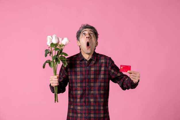 Macho joven de vista frontal sosteniendo hermosas rosas rosadas y tarjeta bancaria en la pared rosa