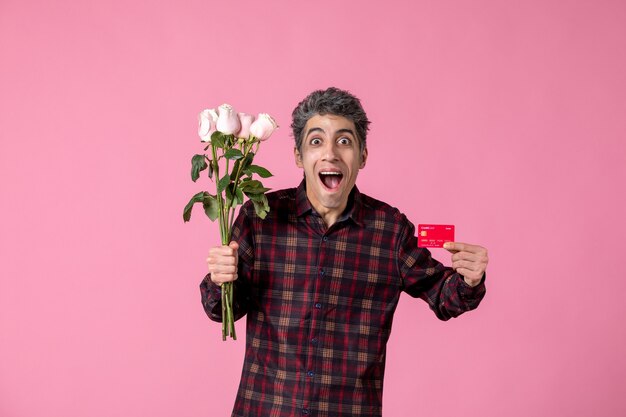 Macho joven de vista frontal sosteniendo hermosas rosas rosadas y tarjeta bancaria en la pared rosa