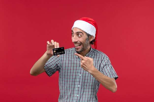 Macho joven de vista frontal sosteniendo una tarjeta bancaria negra en el escritorio rojo