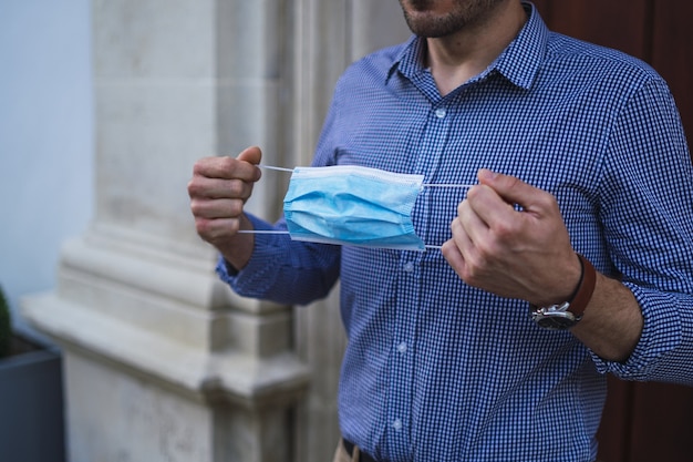 Macho joven vistiendo una camisa azul de pie en la puerta sosteniendo una mascarilla médica