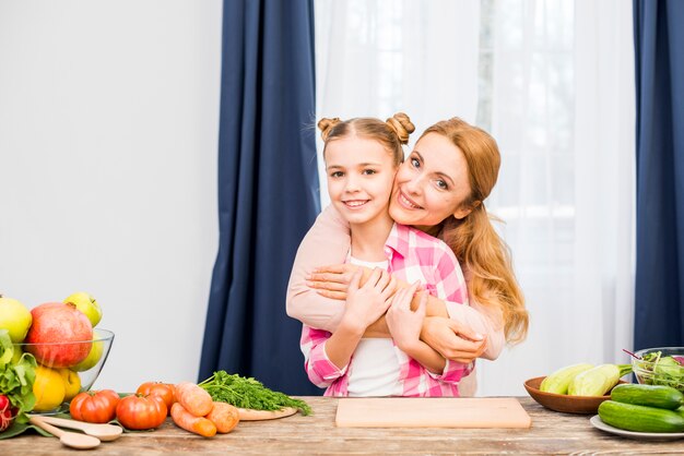 Madre abrazando a su hija de pie detrás de la mesa de madera con verduras frescas