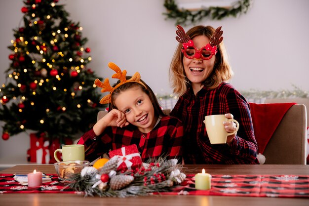 Madre alegre en gafas de reno e hija sentada a la mesa disfrutando de la Navidad en casa