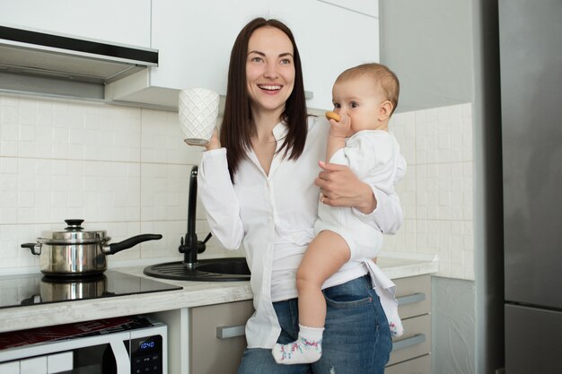 Madre alegre de pie en la cocina, bebiendo café por la mañana y sosteniendo al bebé