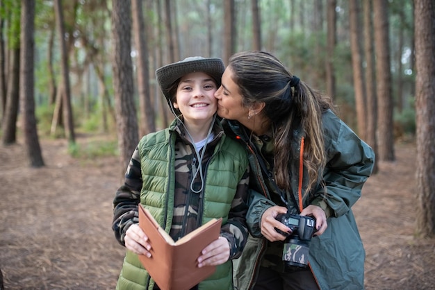 Madre amorosa con hijo con cámara en el bosque. Modelo femenino con ropa deportiva besando a un niño sonriente en la mejilla. Hobby, concepto de fotografía