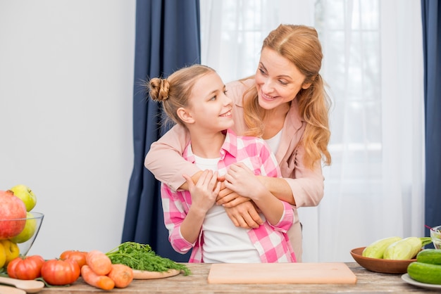 Madre amorosa y su hija de pie detrás de la mesa con verduras