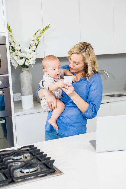 La madre y el bebé que usa el teléfono móvil en la cocina