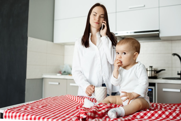 Madre bebiendo café y hablando por teléfono mientras el bebé come en la mesa de la cocina