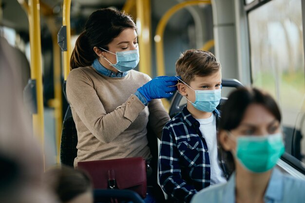 Madre cariñosa poniendo una máscara protectora en la cara de su hijo mientras viaja en autobús