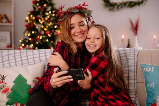 Madre complacida con corona de acebo e hija viendo algo en el teléfono sentado en el sofá y disfrutando de la Navidad en casa