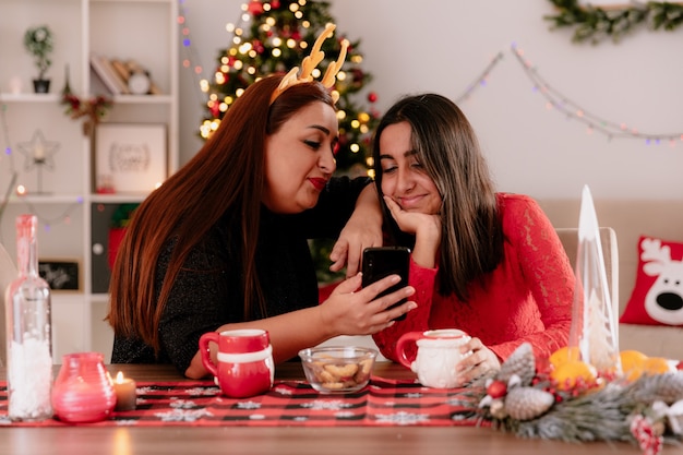 Madre complacida con diadema de renos e hija mire el teléfono sentado a la mesa disfrutando de la Navidad en casa