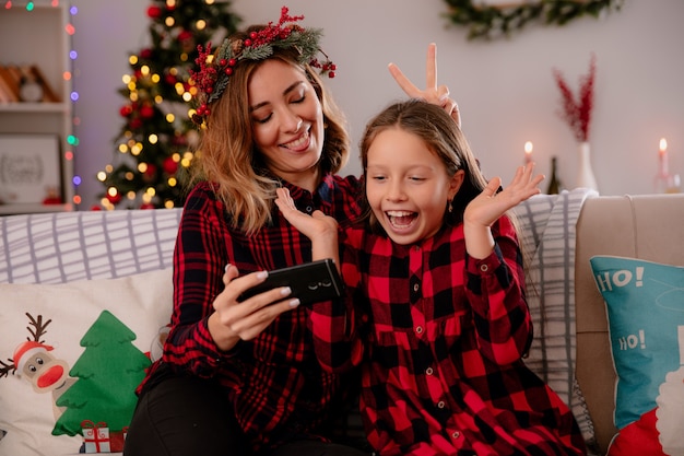 La madre contenta saca la lengua y muestra algo en el teléfono a su hija sentada en el sofá y disfrutando de la Navidad en casa