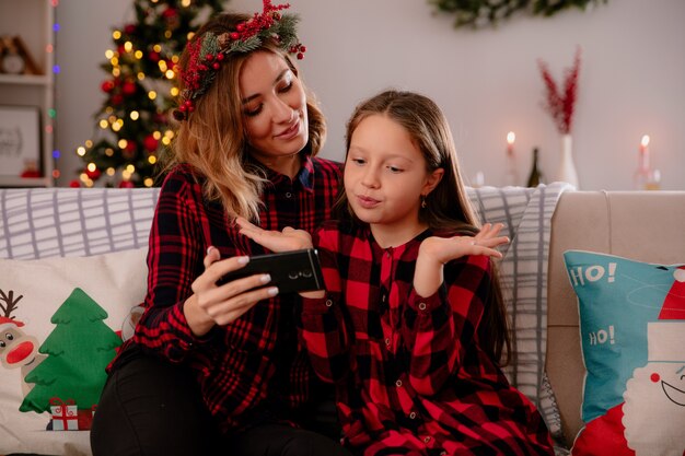 La madre contenta sostiene el teléfono y mira a la hija confundida sentada en el sofá y disfrutando de la Navidad en casa