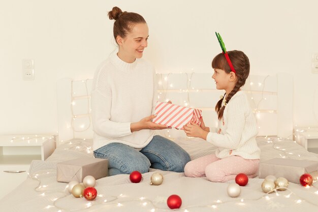 Madre e hija adultas jóvenes vistiendo suéteres blancos de estilo casual sentados en la cama, niña dando regalo de Navidad a su encantadora mamá, feliz año nuevo.